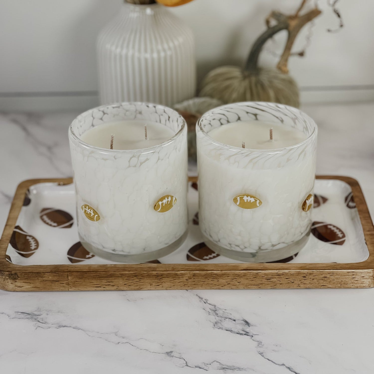 Two white scented candles in clear glass holders with gold football-shaped accents, displayed on a decorative tray with a wood edge. The tray sits on a white marble surface with a white ribbed vase and dried autumn leaves in the background.