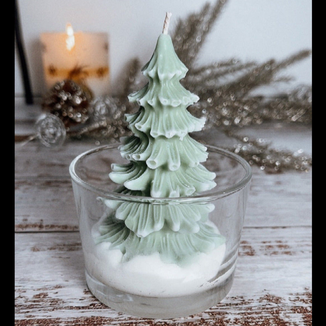A wax candle shaped like a Christmas tree, placed in a clear glass container, with a decorative tree in the background and a lit candle in the top right corner.