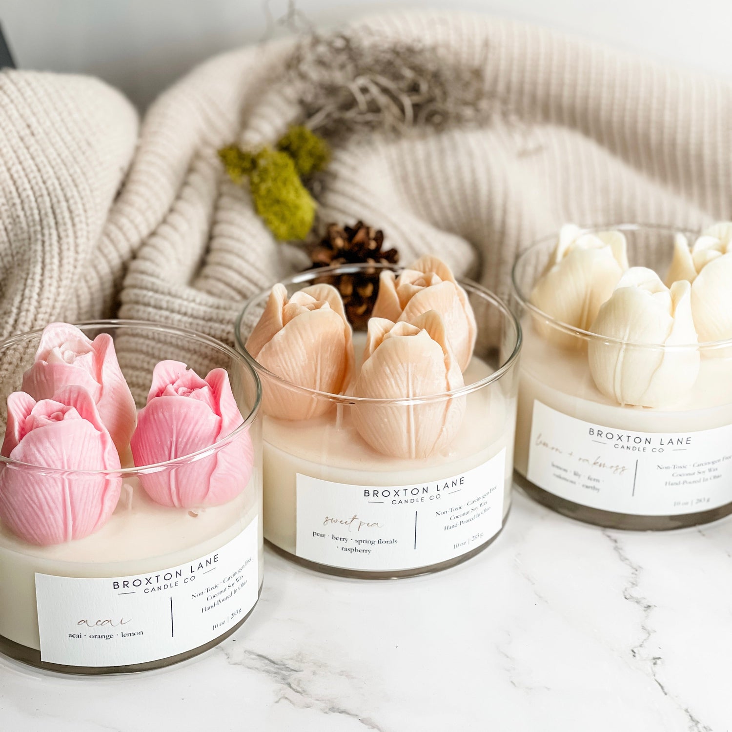 Three tulip-shaped candles in shades of pink and white, set in clear glass jars. The candles are on a marble surface with a beige knit scarf in the background.