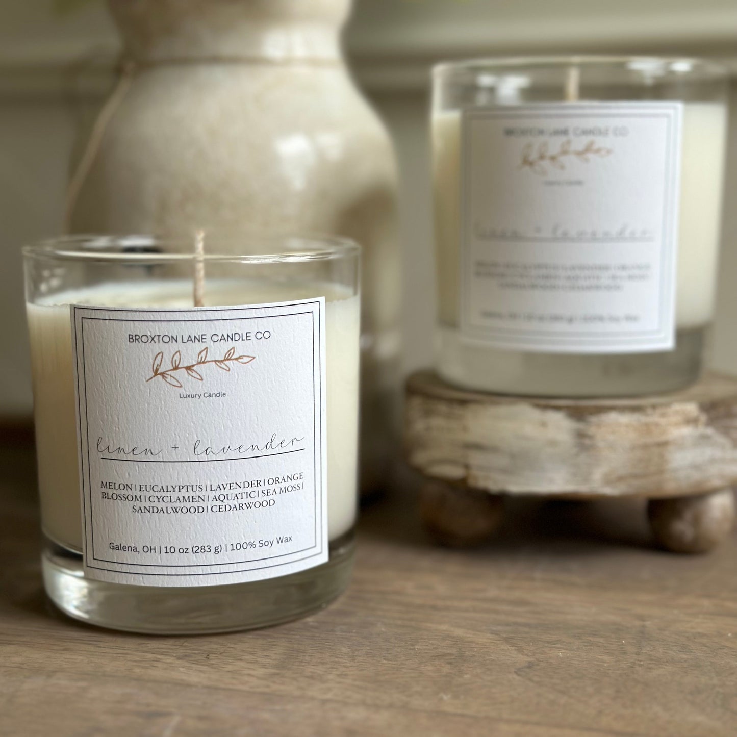 Two clear glass candles with labels, one larger than the other, placed on a wooden surface, with a larger vase containing greenery in the background.