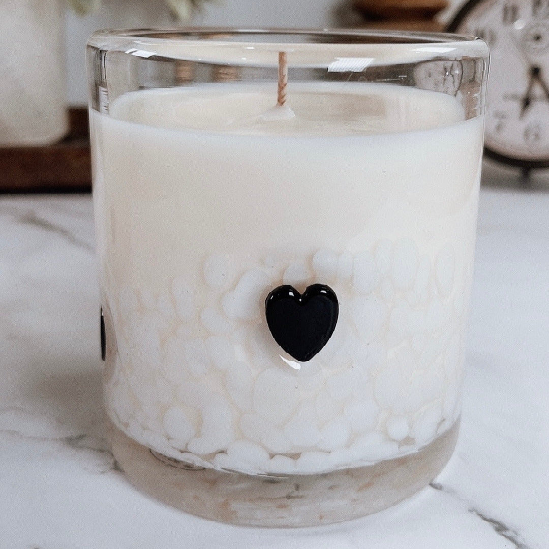 White candle in a glass jar with a black heart-shaped detail on a marble surface.