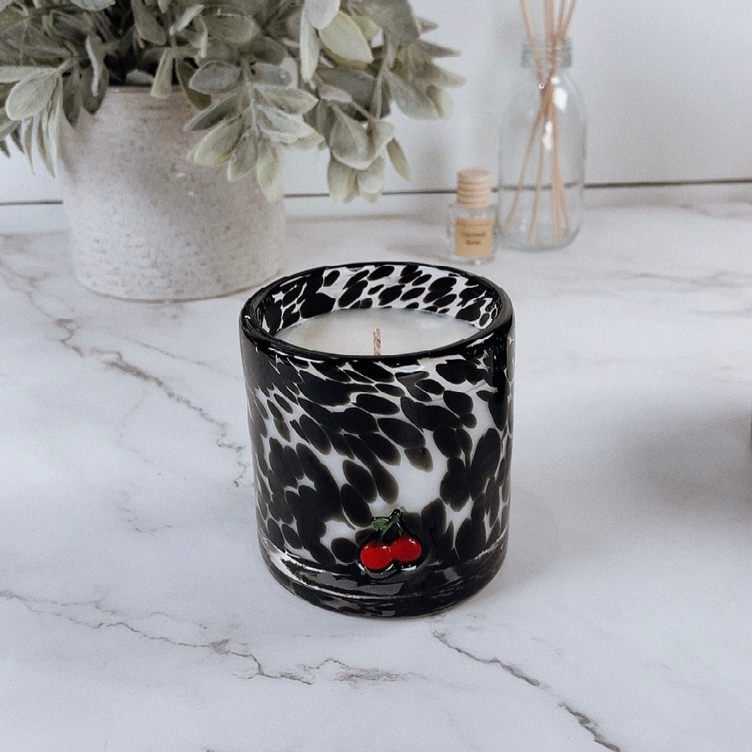 Candle in a black and white patterned glass container on a marble surface with a plant and bottle in the background.