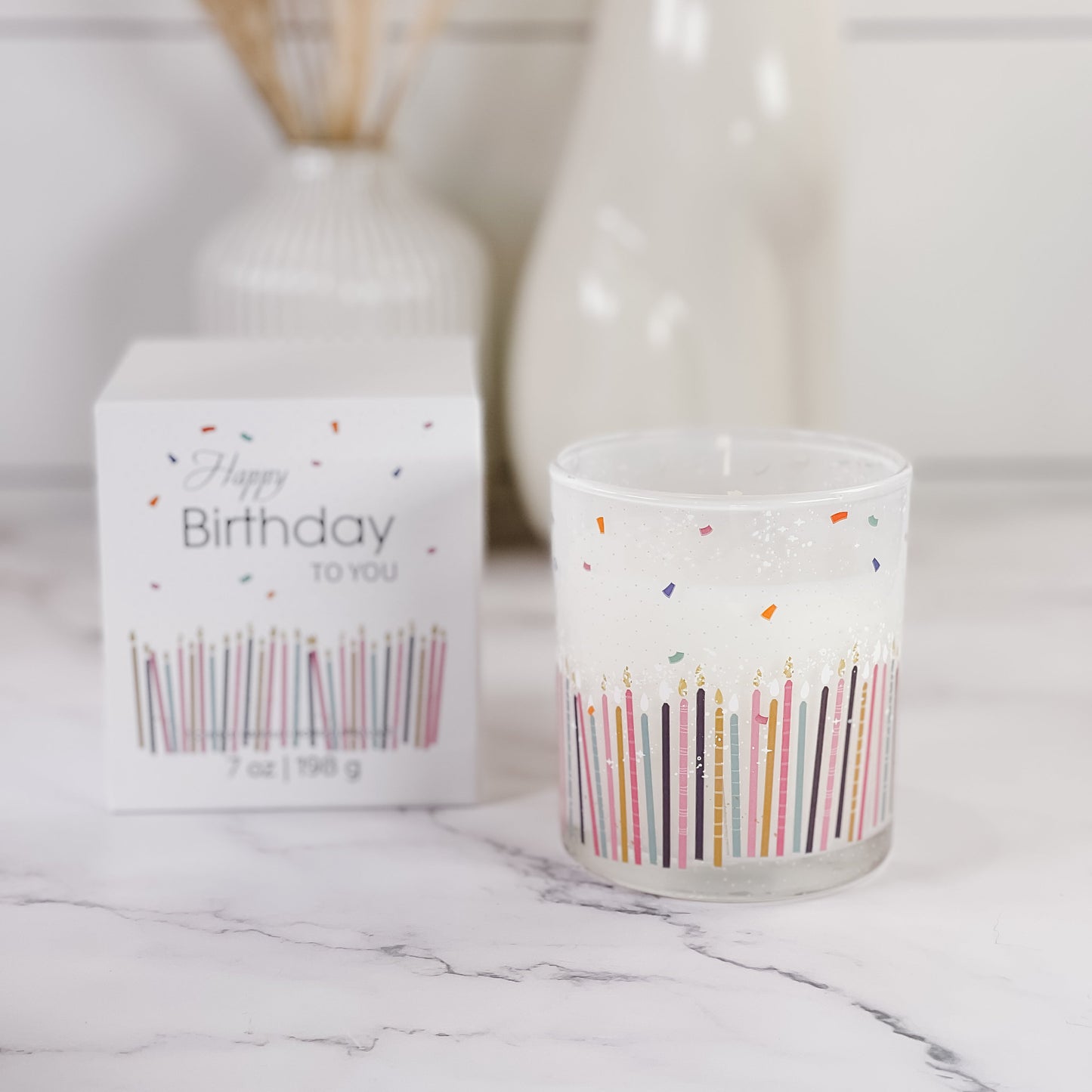 A frosted glass candle jar with colorful birthday candle illustrations sits next to its white 'Happy Birthday to You' box on a white marble surface.
