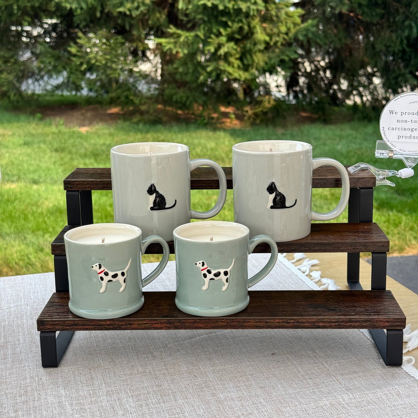 Four ceramic mugs in a pale sage green color with black and white dog illustrations, displayed on a dark wooden stand outdoors.