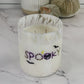 A white wax candle in a clear glass jar with a white marbled rim, decorated with the word 'spooky' in purple and black with small bat and pumpkin motifs. The candle is set on a white marble surface with decorative pumpkins and a ribbed vase in the background.