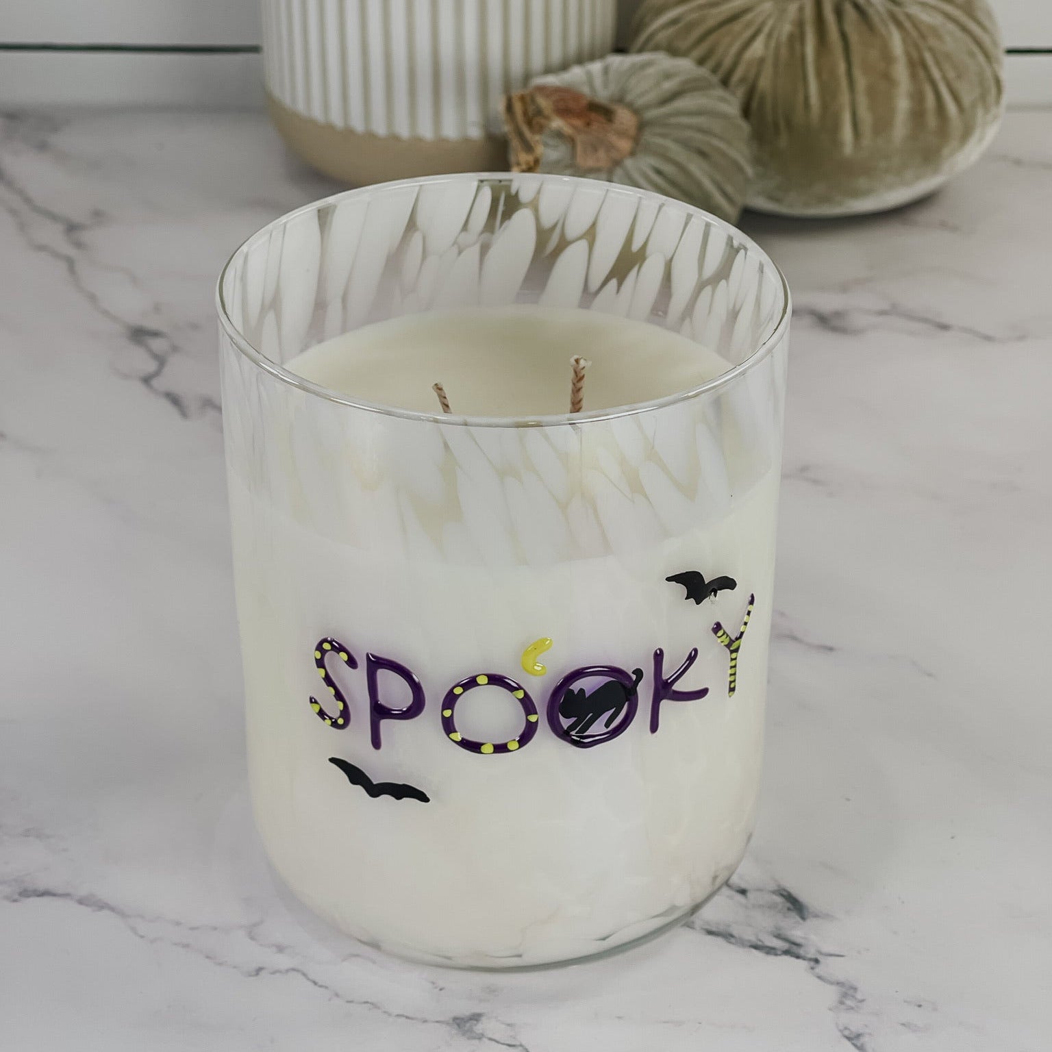 A white wax candle in a clear glass jar with a white marbled rim, decorated with the word 'spooky' in purple and black with small bat and pumpkin motifs. The candle is set on a white marble surface with decorative pumpkins and a ribbed vase in the background.