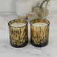 Two leopard-print glass candle holders with white wax candles, set on a white marble surface with a blurred background of a vase and dried flowers.