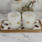 Two white scented candles in clear glass holders with gold football-shaped accents, displayed on a decorative tray with a wood edge. The tray sits on a white marble surface with a white ribbed vase and dried autumn leaves in the background.