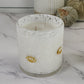 A white scented candle in a clear glass jar with a white marbled pattern and small gold football-shaped accents, set on a white marble surface with decorative pumpkins and a ribbed ceramic vase in the background.