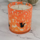 An orange and white speckled glass candle jar with a small black cat silhouette on the front, set on a white marble surface with decorative pumpkins in the background.