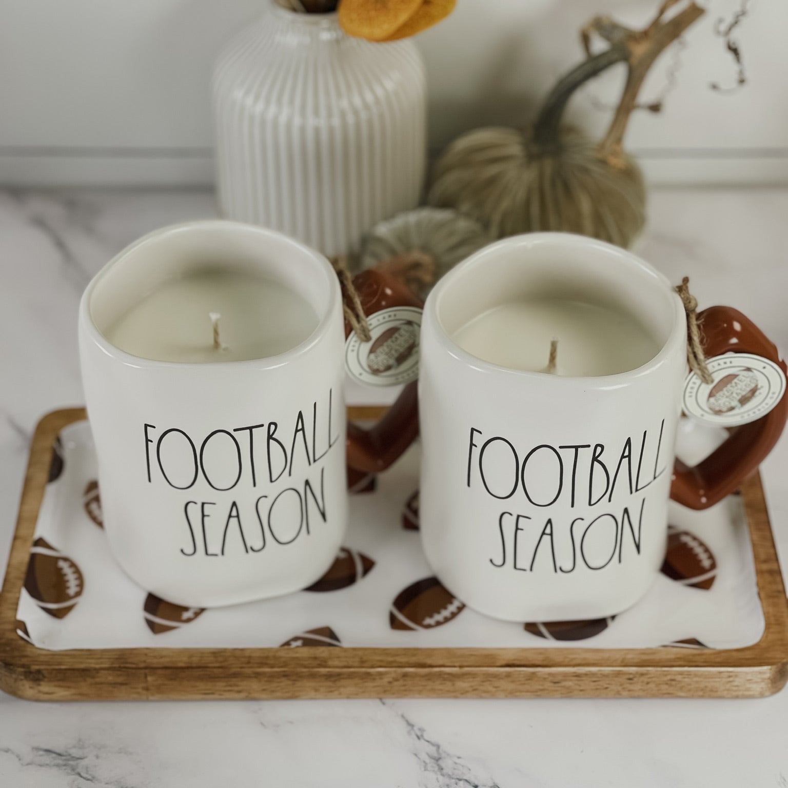 Two white ceramic mugs with brown handles, each printed with 'FOOTBALL SEASON' in black text, displayed on a wooden tray with football-shaped linens. Decorative pumpkins and a white ribbed vase with dried leaves sit in the background on a white marble surface.