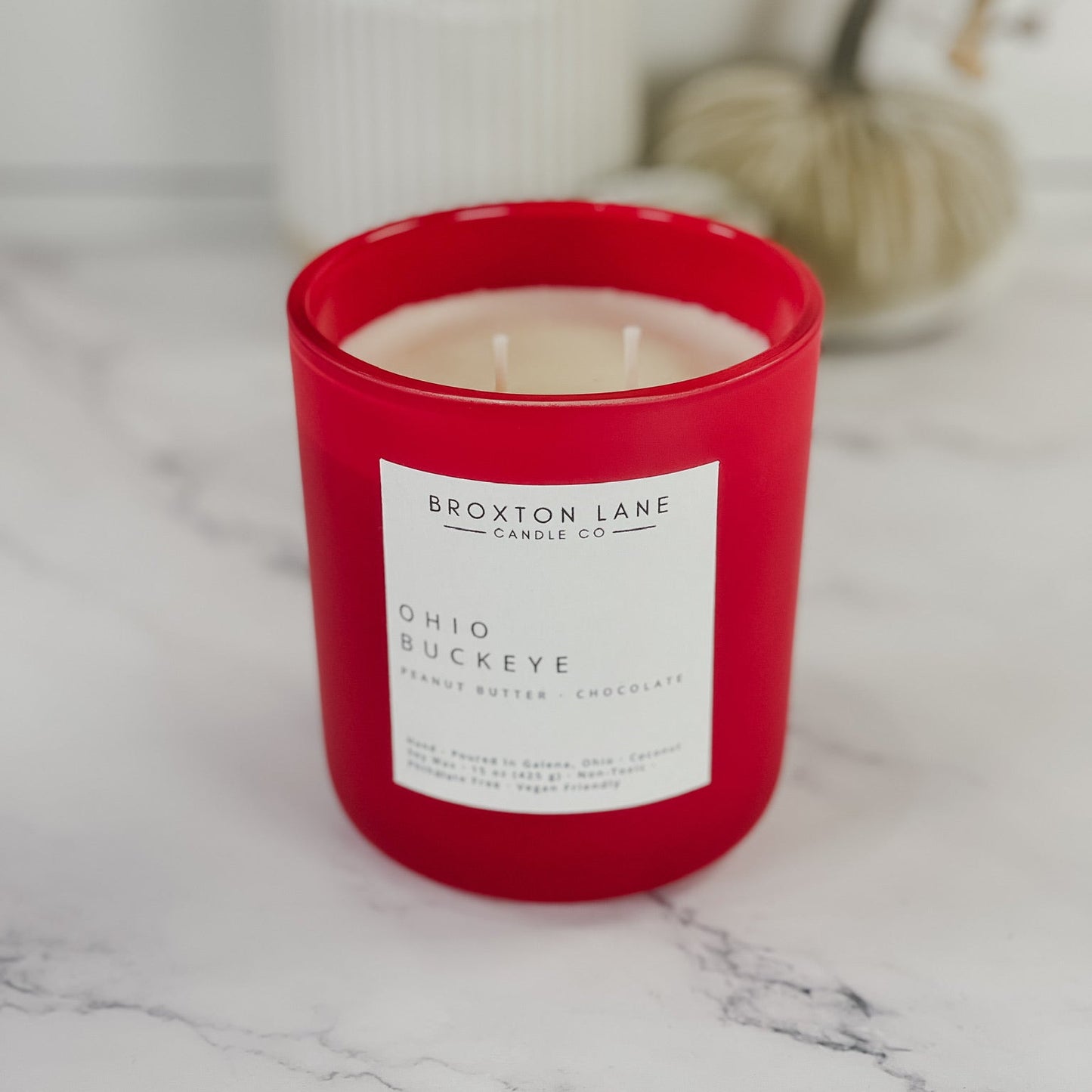 A red glass jar candle with a white label reading 'Brooklyn Lane Candle Co. Ohio Buckeye' sits on a white marble surface with soft-focus decorative pumpkins in the background.