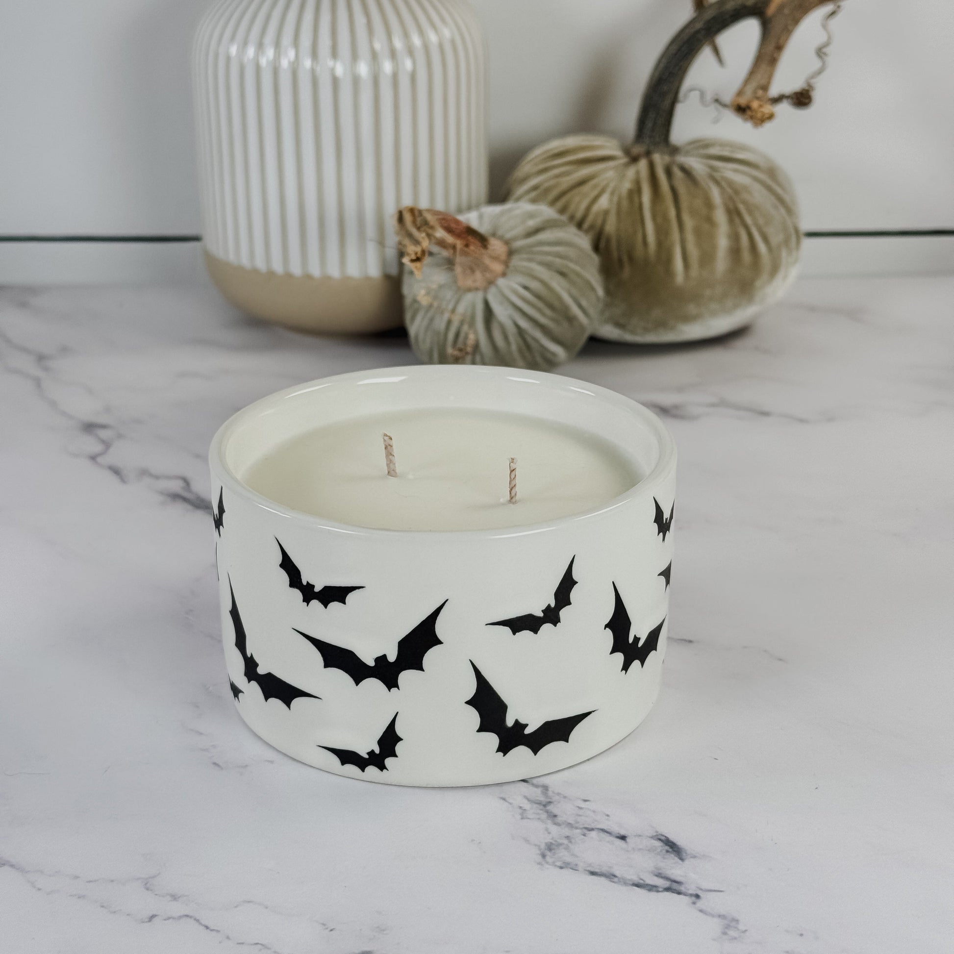 A white ceramic two-wick candle with black bat silhouettes printed around the outside, styled on a white marble surface with decorative pumpkins and a ribbed vase in the background.