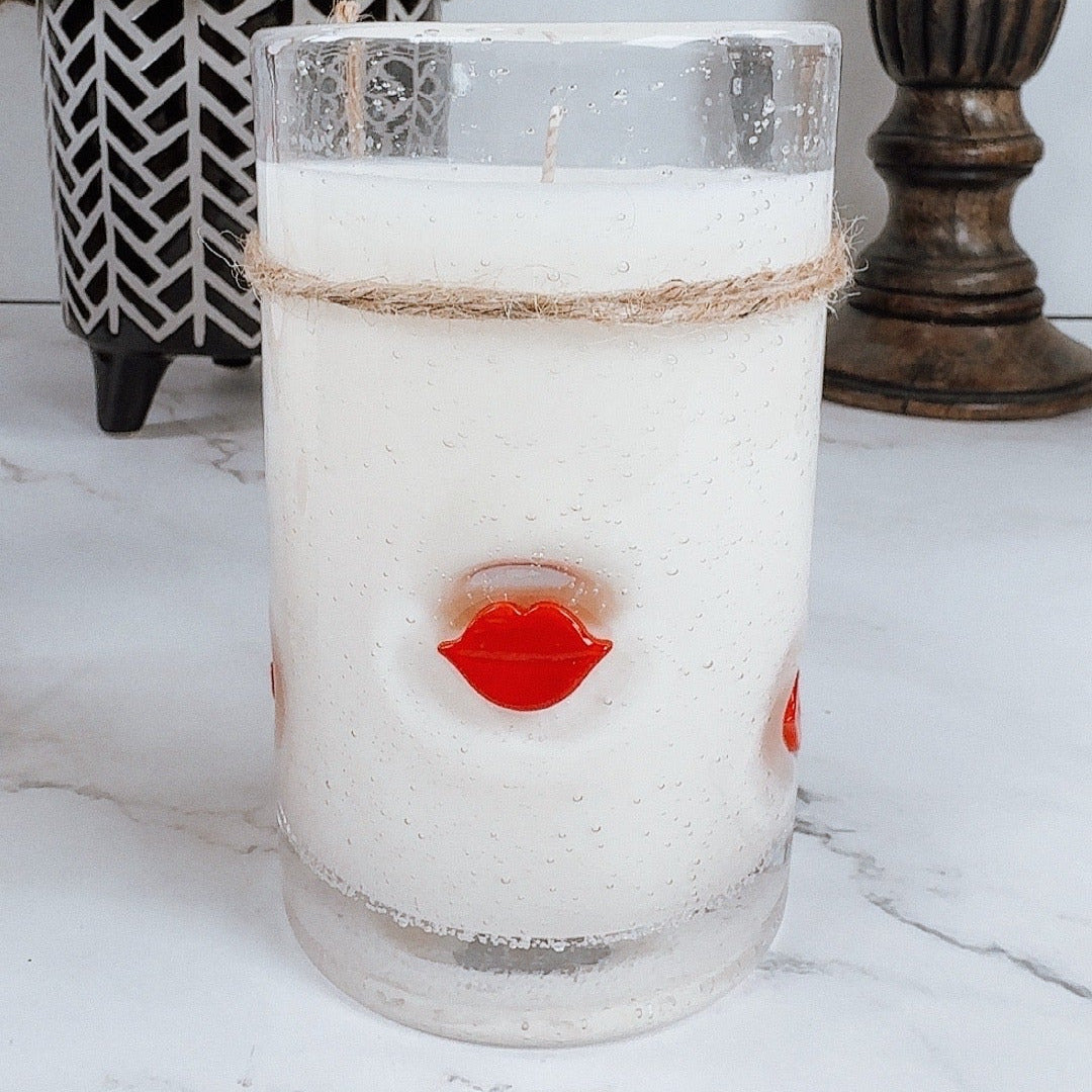 Candle in a glass jar with red lips on a white surface, with decorative elements in the background.