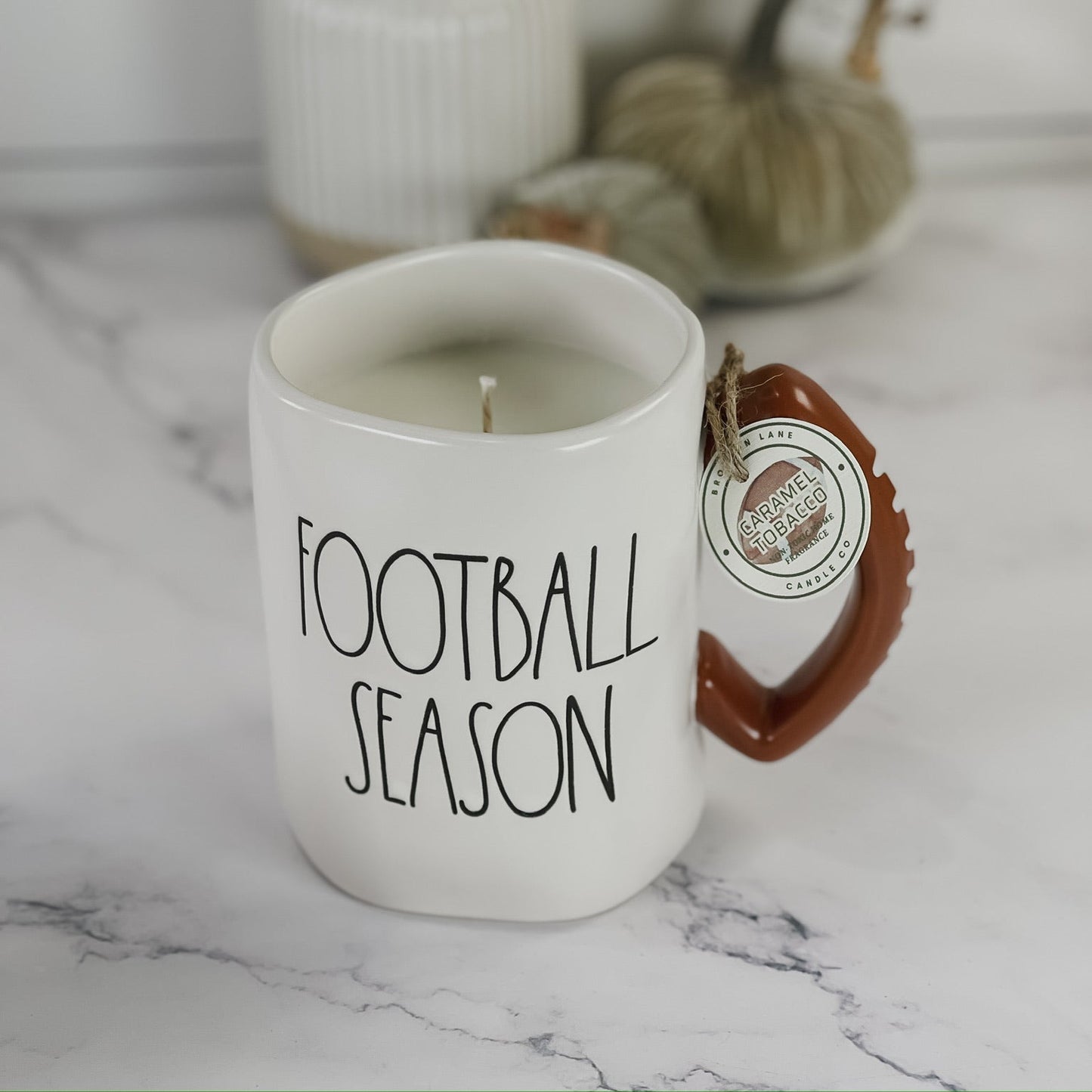 A white ceramic candle mug with 'FOOTBALL SEASON' printed in black text, featuring a brown handle and a hanging circular tag. The mug is styled on a white marble surface with a white ribbed vase and dried yellow leaves in the background.
