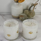 Two white scented candles in textured clear glass jars with small gold football-shaped labels, styled on a marble surface with dried flowers and pumpkins in the background.