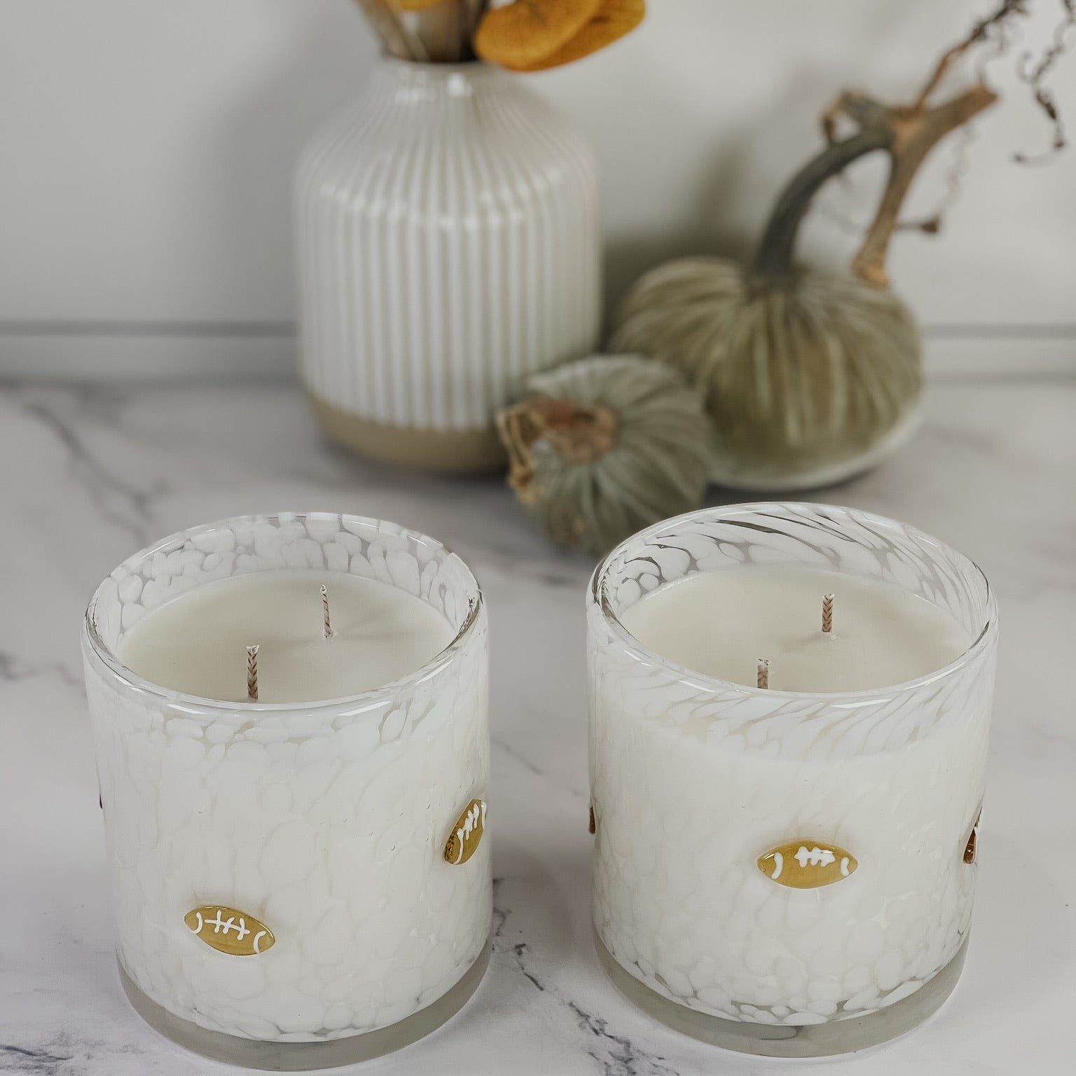 Two white scented candles in textured clear glass jars with small gold football-shaped labels, styled on a marble surface with dried flowers and pumpkins in the background.