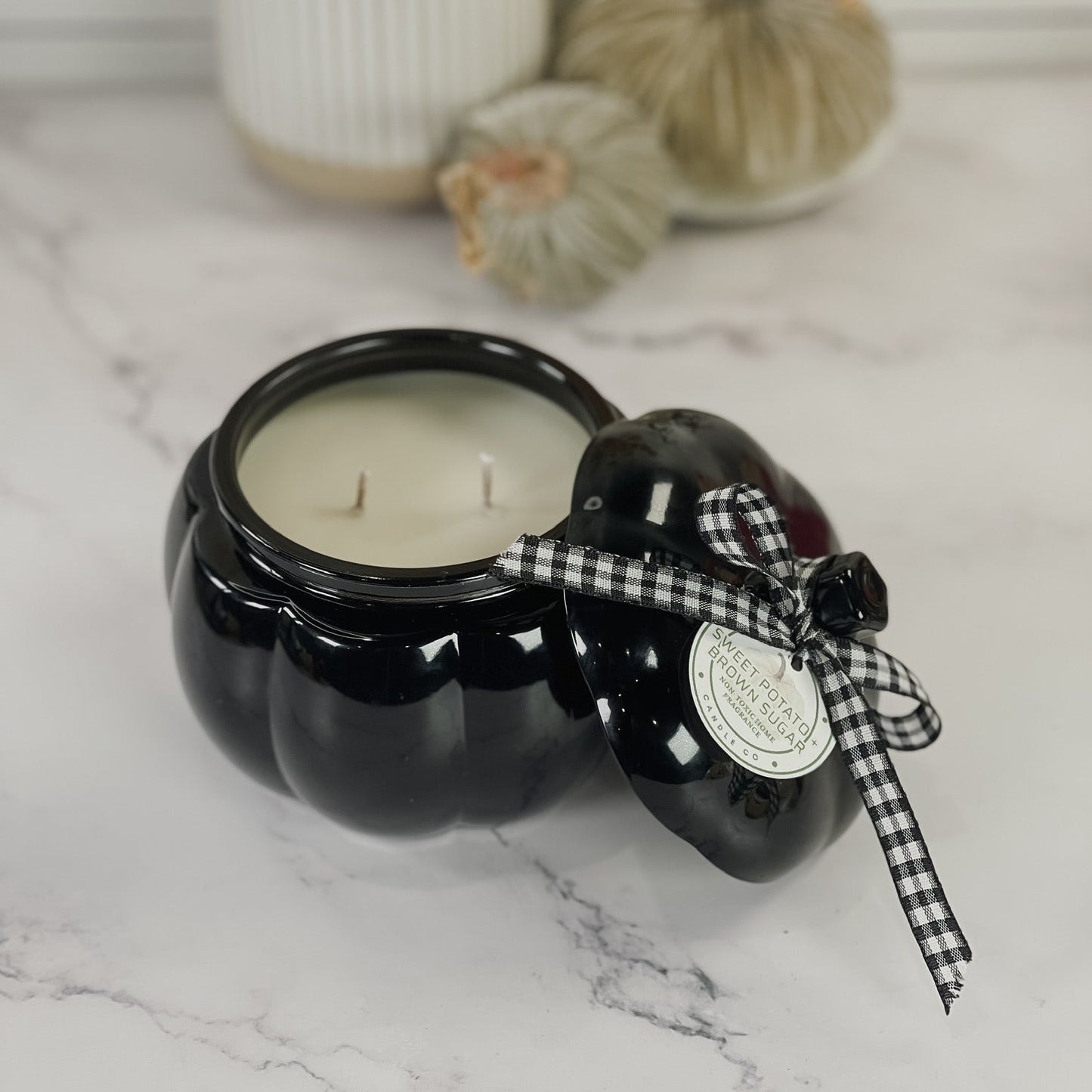 A black pumpkin-shaped candle jar with a matching lid, tied with a black-and-white gingham ribbon and a round paper seal, set on a white marble surface with decorative pumpkins and a ribbed vase in the background.