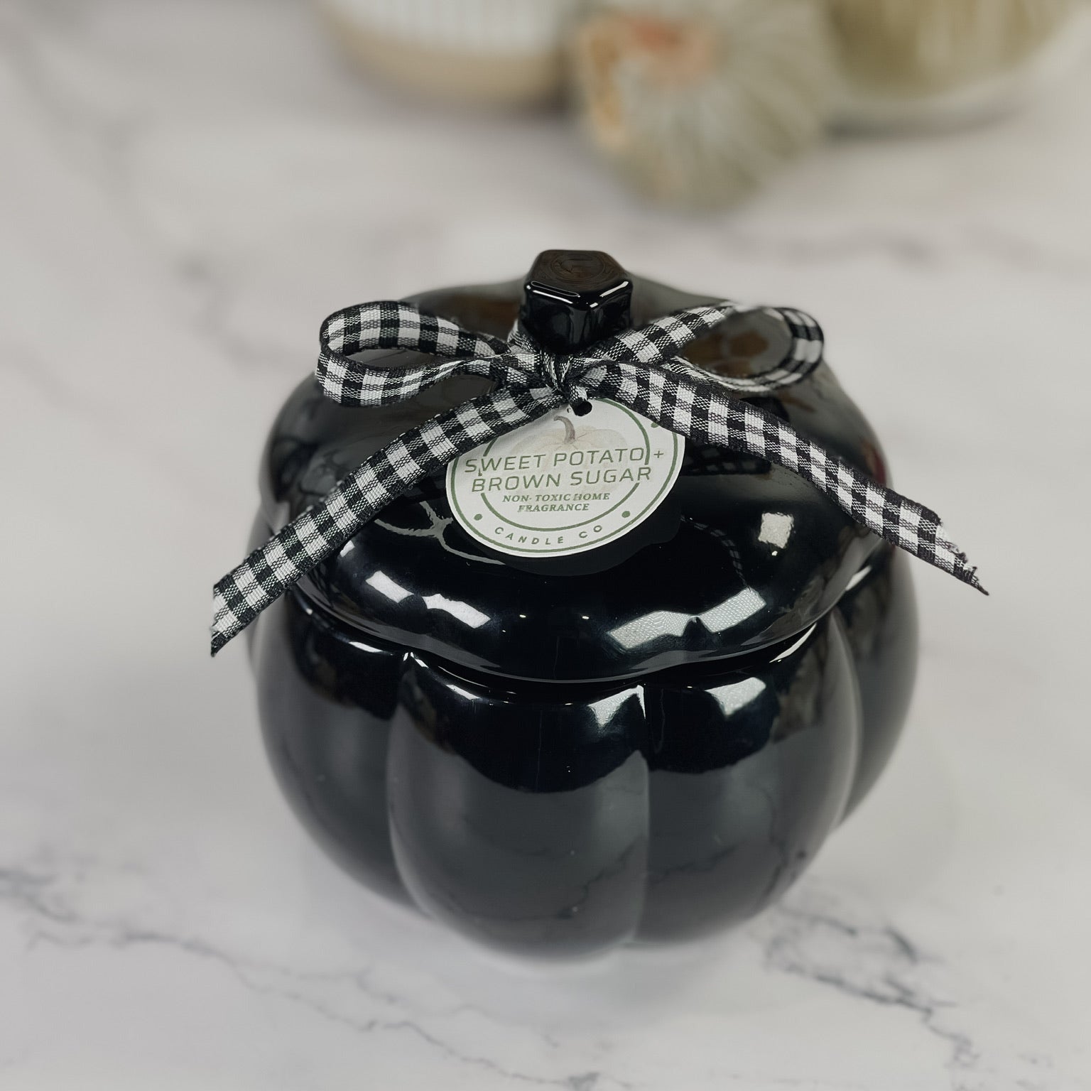 A glossy black pumpkin-shaped lidded container with a black-and-white gingham ribbon bow on top, featuring a round white label that reads 'SWEET POTATO BROWN SUGAR'. The container is placed on a white marble surface with a softly blurred background containing neutral decor.