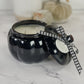 A black pumpkin-shaped candle jar with a matching lid tied in a black-and-white gingham ribbon, set on a white marble surface.