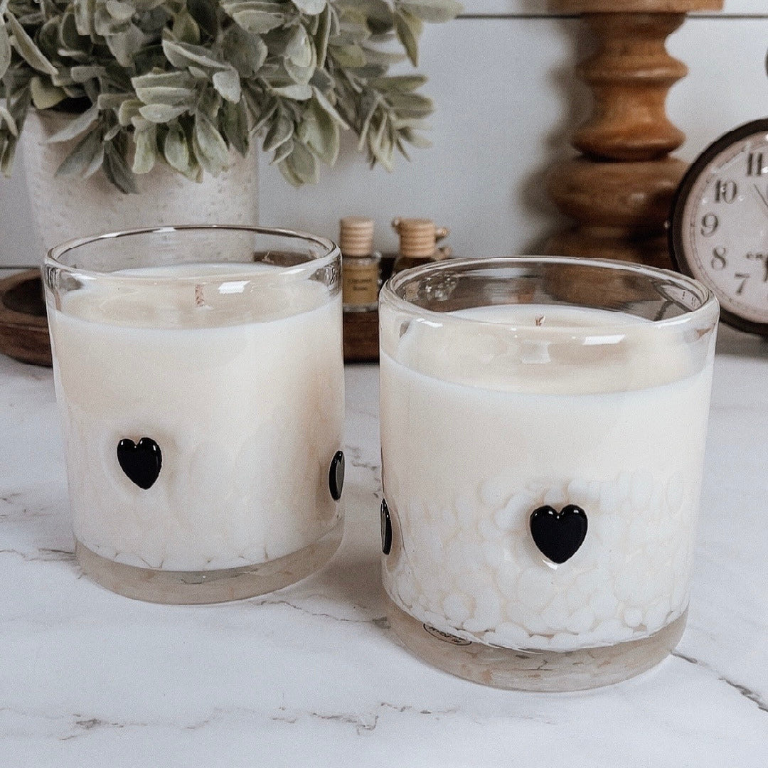 Two glass candles with black heart details on a marble surface, with a plant and clock in the background.