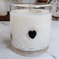 White candle in a glass jar with a black heart-shaped detail on a marble surface.
