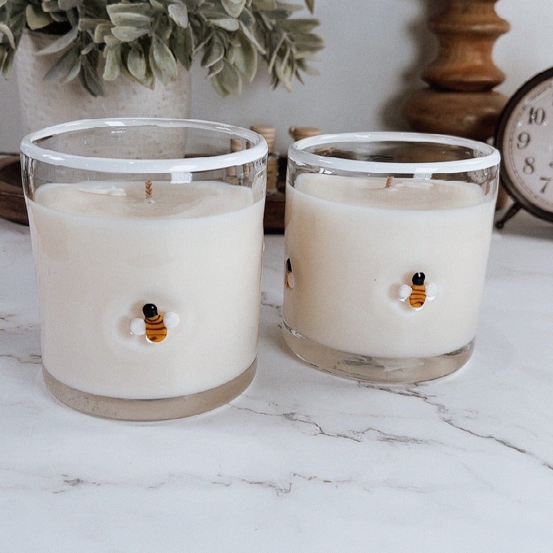 Two candles with bees decorations in glass holders on a marble surface with plants and a clock in the background.