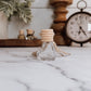 Small glass bottle with wooden cap on a marble surface, with decorative items in the background.