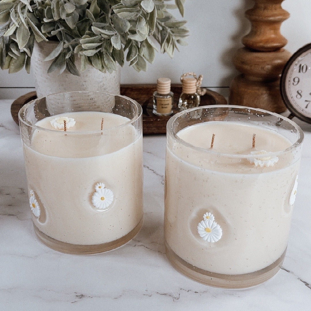 Two candles with floral designs on a white surface with a plant and wooden items in the background.