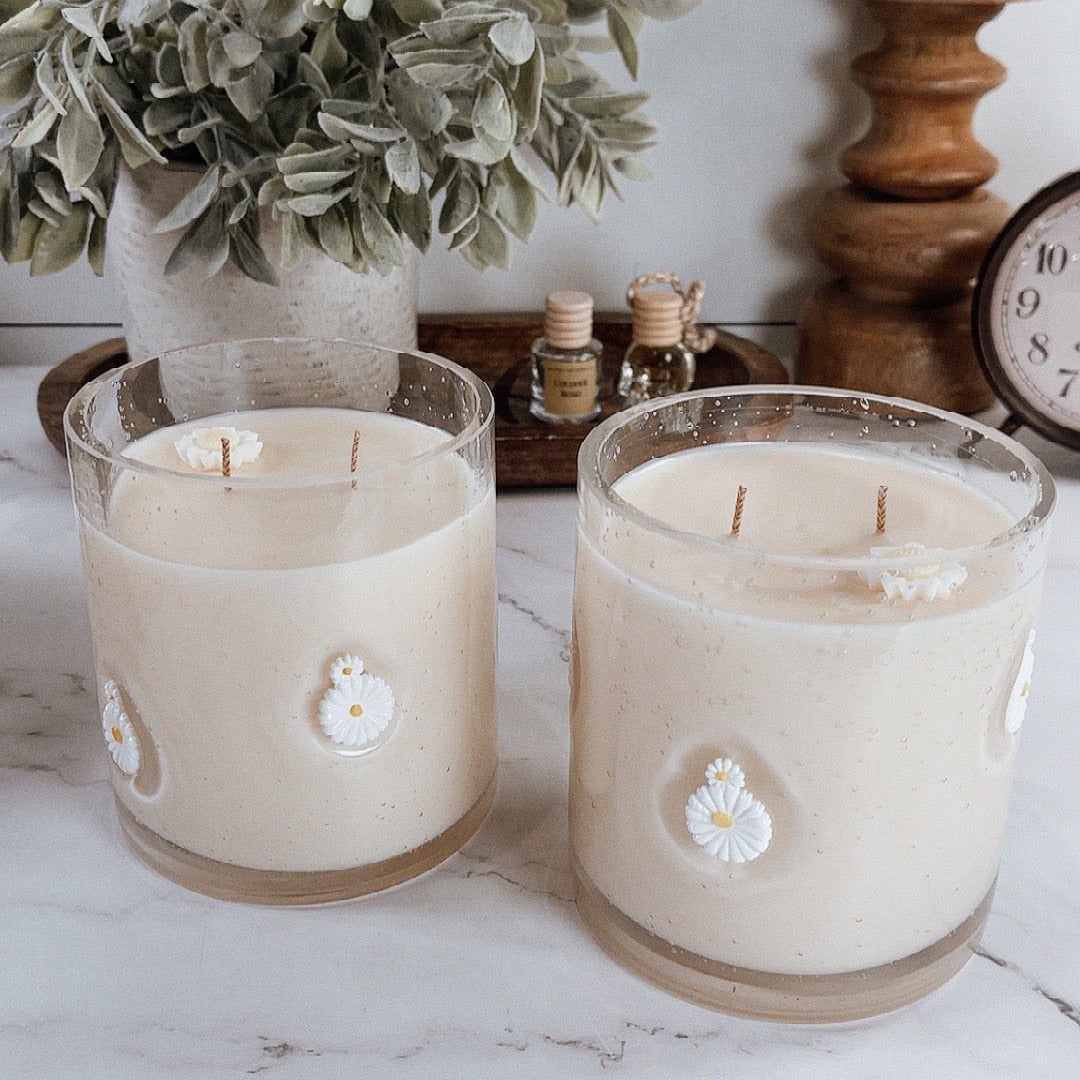 Two candles with floral designs on a white surface with a plant and wooden items in the background.