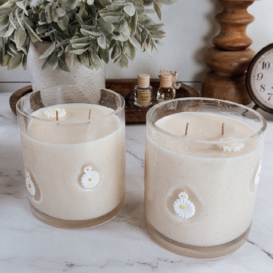 Two candles with floral designs on a white surface with a plant and wooden items in the background.