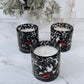 Three black and white patterned candles with red accents on a marble surface, with plants in the background.