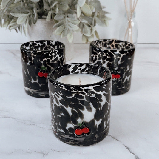 Candle in a leopard print container with decorative cherries on a marble surface, surrounded by plants.