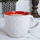 Speckled ceramic mug with candle inside  with red interior on a marble surface, next to a potted plant and decorative item.