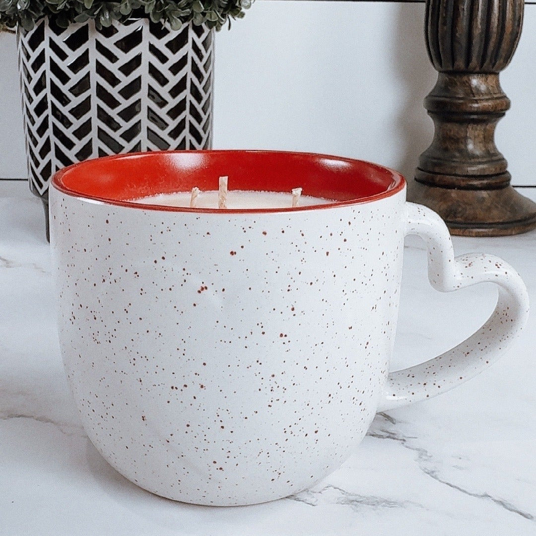Speckled ceramic mug with candle inside  with red interior on a marble surface, next to a potted plant and decorative item.