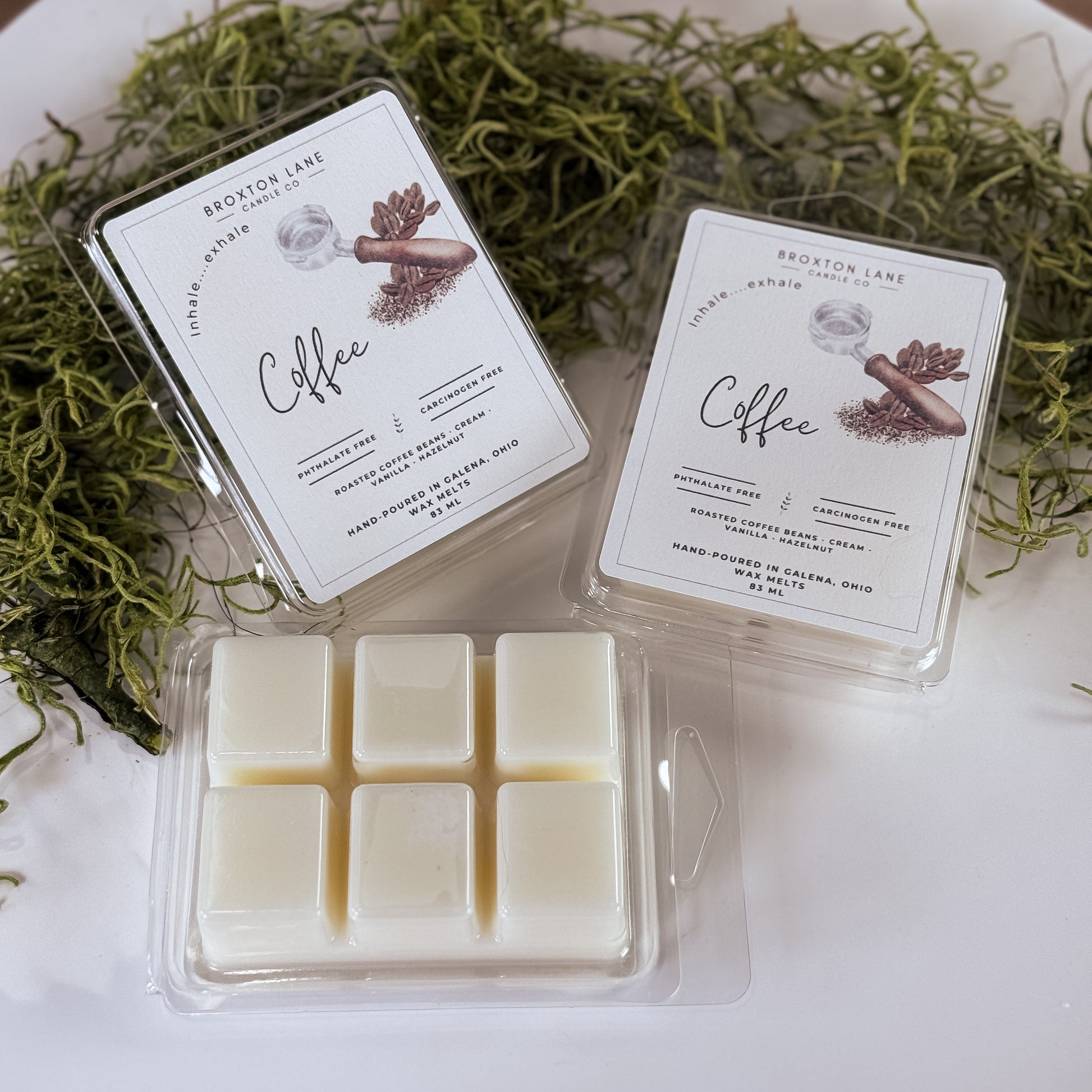 Two packages of coffee-scented wax melts on a white surface with greenery.