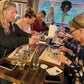 Group of people gathered around a table with wine glasses, in a cozy indoor setting.