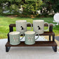 Four ceramic mugs in a pale sage green color with black and white dog illustrations, displayed on a dark wooden stand outdoors.